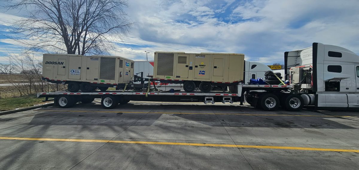 Two large portable air compressors strapped down on a step deck trailer
